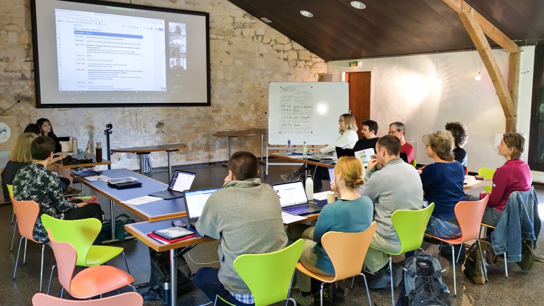 Several people sitting around a u-shaped table and looking at a white screen on the wall in a meeting room at the pause-and-reflect workshop