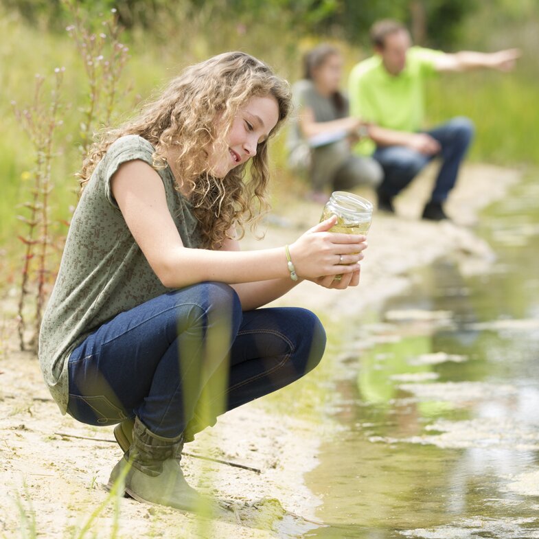 Smiling child looking at a jar with a sample, squatting next on the shoreline, perhaps doing a pause and reflect on the data in the jar